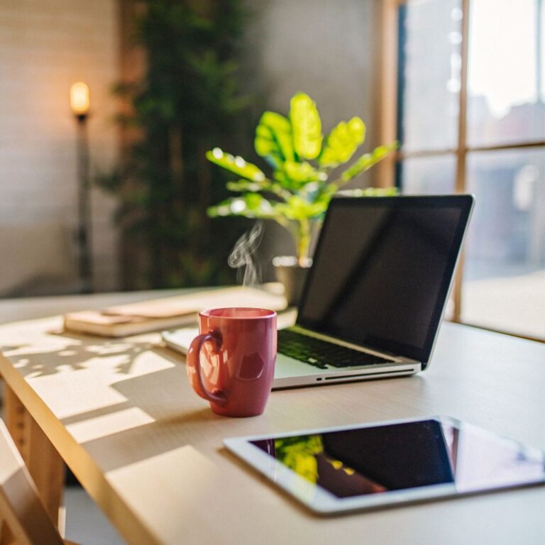 A laptop, tablet and coffee mug can be seen on a desk in front of a window.