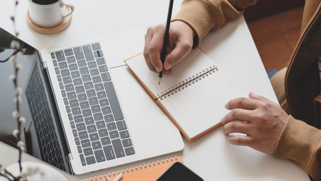 Shows a pair of hands writing in a blank notebook at a computer desk.